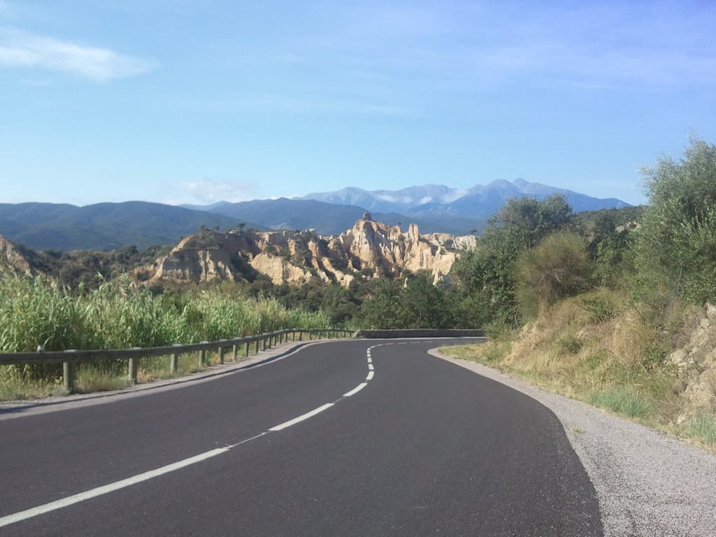 <p>they're called Les Orgues d'Ille-sur-T&ecirc;t. &nbsp;From here the descent steepens and becomes quite curvy, I could have gone faster but had a camper van in front of me that I couldn't overtake without taking risks.</p><p>After Ille-sur-T&ecirc;t it was flat or even downhill to Thuir but the wind was very strong, at various points I was pedalling hard in bottom gear going downhill! &nbsp;Very depressing.</p><p>Thuir was good in that I found an epicerie that was doing freshly roasted chickens, and they'd sell me half a one. &nbsp;So, fighting the wind that was threatening to blow my chicken away, I ate it and half a baguette in the shade of a tree just on the outskirts.</p>
